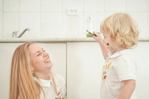 A cheerful mother and a joyful child sit on the floor in white t-shirts and paint each other with paints — Stock Photo, Image