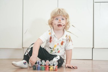 İki yaşında, elleri ve yüzü renkli boyalarla boyanmış daha fazla eğlenceye hazır bir çocuk. Kirli ve mutlu çocuk beyaz oda arka planında yerde oturuyor..