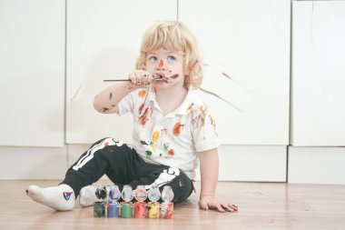 İki yaşında, elleri ve yüzü renkli boyalarla boyanmış daha fazla eğlenceye hazır bir çocuk. Kirli ve mutlu çocuk beyaz oda arka planında yerde oturuyor..