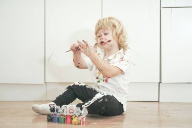 İki yaşında, elleri ve yüzü renkli boyalarla boyanmış daha fazla eğlenceye hazır bir çocuk. Kirli ve mutlu çocuk beyaz oda arka planında yerde oturuyor..