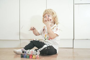 İki yaşında, elleri ve yüzü renkli boyalarla boyanmış daha fazla eğlenceye hazır bir çocuk. Kirli ve mutlu çocuk beyaz oda arka planında yerde oturuyor..