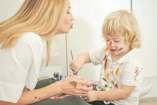 A cheerful mother and a joyful child sit on the floor in white t-shirts and paint each other with paints — Stock Photo, Image