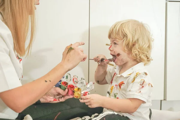 A cheerful mother and a joyful child sit on the floor in white t-shirts and paint each other with paints — Stock Photo, Image