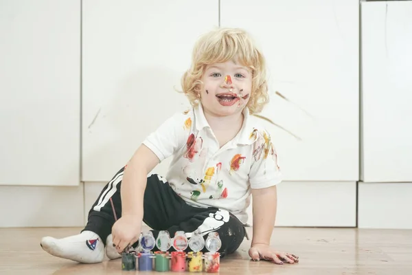 Δίχρονο αγόρι με τα χέρια και το πρόσωπο βαμμένα σε πολύχρωμα χρώματα έτοιμα για περισσότερη διασκέδαση. βρώμικο και χαρούμενο παιδί που κάθεται στο πάτωμα σε λευκό φόντο δωματίου. — Φωτογραφία Αρχείου