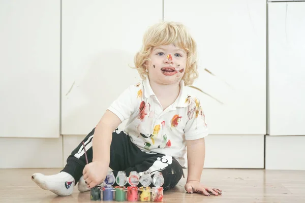 Δίχρονο αγόρι με τα χέρια και το πρόσωπο βαμμένα σε πολύχρωμα χρώματα έτοιμα για περισσότερη διασκέδαση. βρώμικο και χαρούμενο παιδί που κάθεται στο πάτωμα σε λευκό φόντο δωματίου. — Φωτογραφία Αρχείου