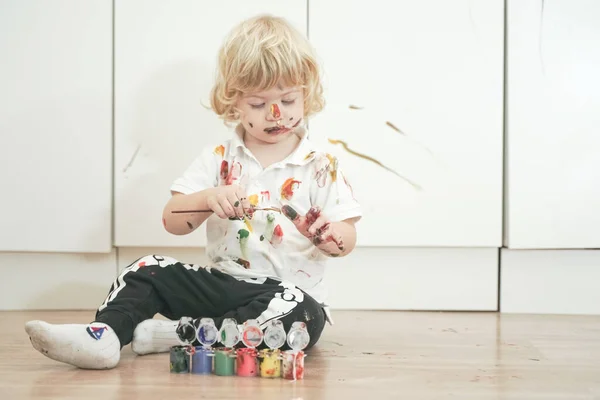 Menino de dois anos com as mãos e rosto pintado em tintas coloridas pronto para mais diversão. criança suja e feliz sentado no chão no fundo da sala branca . — Fotografia de Stock