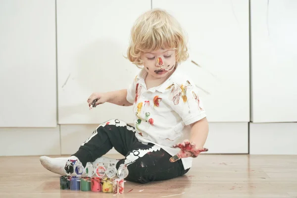 Niño de dos años con las manos y la cara pintada en pinturas de colores listos para más diversión. niño sucio y feliz sentado en el suelo en el fondo de la habitación blanca . — Foto de Stock