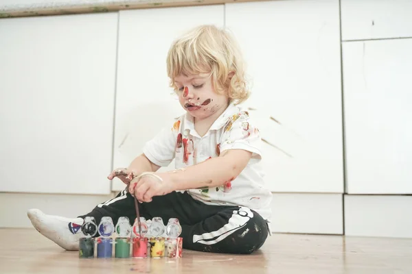 Two year old boy with hands and face painted in colorful paints ready for more fun. dirty and happy kid sitting on the floor in white room background. — Stock Photo, Image