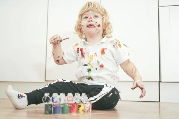 Två år gammal pojke med händer och ansikte målade i färgglada färger redo för roligare. smutsiga och glada barn sitter på golvet i vitt rum bakgrund. — Stockfoto