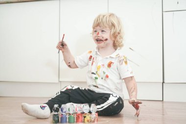 İki yaşında, elleri ve yüzü renkli boyalarla boyanmış daha fazla eğlenceye hazır bir çocuk. Kirli ve mutlu çocuk beyaz oda arka planında yerde oturuyor..