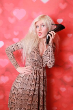 Genç bir kadının ayakkabıyı yüzüne yakın tutan bir telefon gibi kullanması ve konuşması, kırmızı arka plan. Pin-up tarzı.