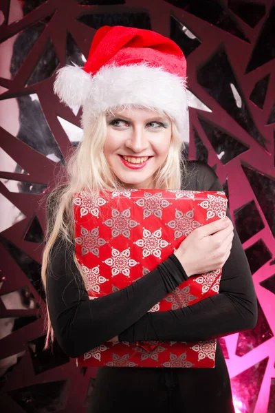 Счастливая милая стройная молодая женщина в рождественской шляпе с подарочной коробкой на темном фоне ночного клуба. плохая игривая девочка санта . — стоковое фото