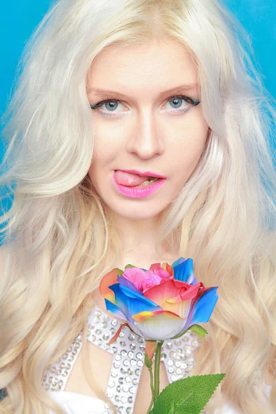 Милая игривая блондинка в белом платье стоит с экологически чистой искусственной розы на синем фоне в студии — стоковое фото
