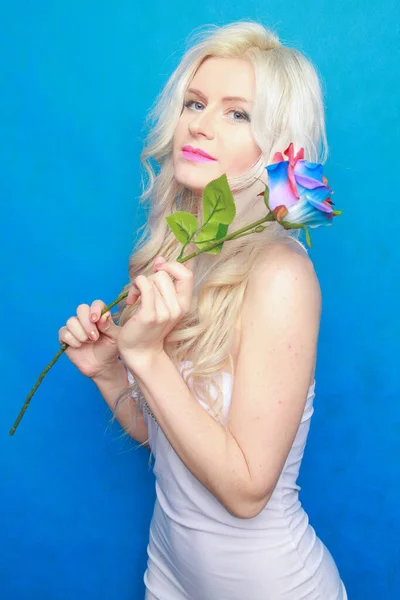 Carino giocoso ragazza bionda in abito bianco stand con eco friendly rosa artificiale su sfondo blu in Studio — Foto Stock