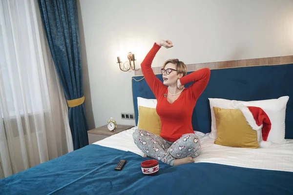 Schattig plus size kort haar blond meisje in bril gaapt in de ochtend in bed zitten in een pyjama en stretching — Stockfoto