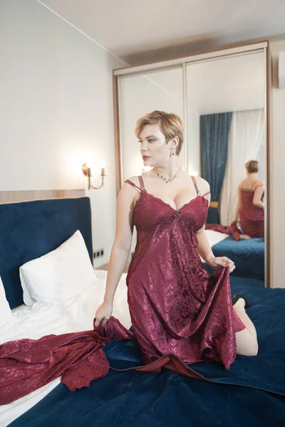 Elegante pin up pelo corto mujer rubia con cuerpo curvilíneo de tamaño grande posando en albornoz rojo de moda en el dormitorio solo — Foto de Stock