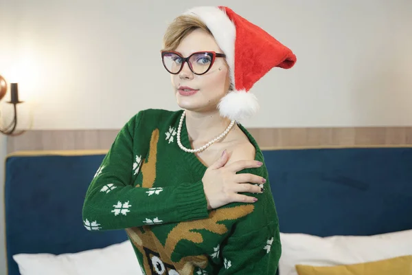 Cute plus rozmiar nerd dziewczyna w okularach i zielony sweter świąteczny z reniferów i czerwony kapelusz Świętego Mikołaja rano na łóżku pozowanie — Zdjęcie stockowe