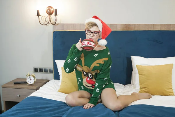 Mignon plus la taille nerd fille dans des lunettes et vert pull de Noël avec rennes et rouge chapeau du Père Noël le matin sur le lit posant — Photo