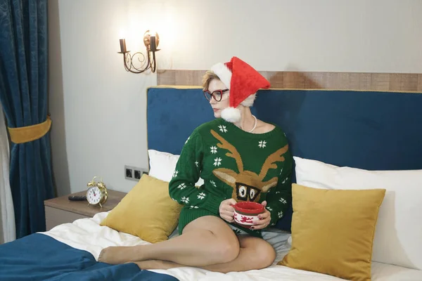 Mignon plus la taille nerd fille dans des lunettes et vert pull de Noël avec rennes et rouge chapeau du Père Noël le matin sur le lit posant — Photo