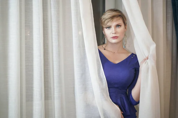 Hermosa rubia más tamaño joven en vestido azul de pie junto a la ventana cerca de cortinas blancas — Foto de Stock