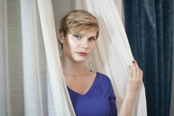 Beautiful blonde plus size young woman in blue dress standing by the window near white curtains