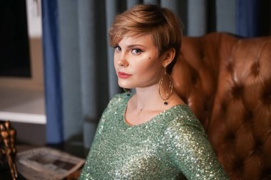 Çekici kısa saçlı, kıvrımlı vücutlu, gece dar elbiseli, deri koltukta oturan sarışın kız.