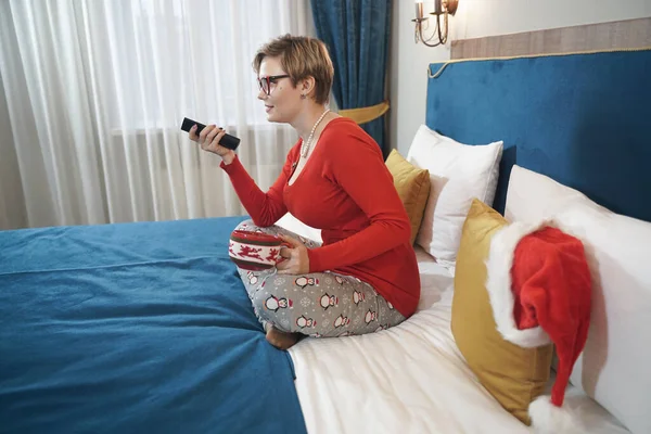 Νεαρή γυναίκα με κόκκινο χριστουγεννιάτικο πουλόβερ βλέποντας τηλεόραση στο κρεβάτι — Φωτογραφία Αρχείου