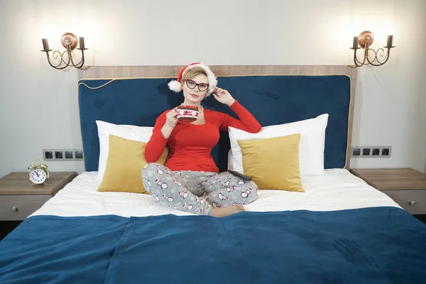 Νεαρή γυναίκα με κόκκινο χριστουγεννιάτικο πουλόβερ βλέποντας τηλεόραση στο κρεβάτι — Φωτογραφία Αρχείου