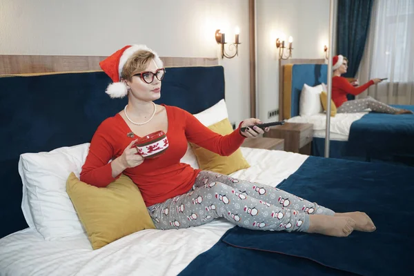 Młoda kobieta w czerwonym swetrze świątecznym ogląda telewizję na łóżku — Zdjęcie stockowe