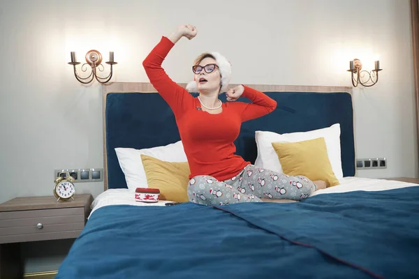 Сонная женщина в красном свитере и шляпе Санты растягивается, вставая утром после Рождества на кровати — стоковое фото