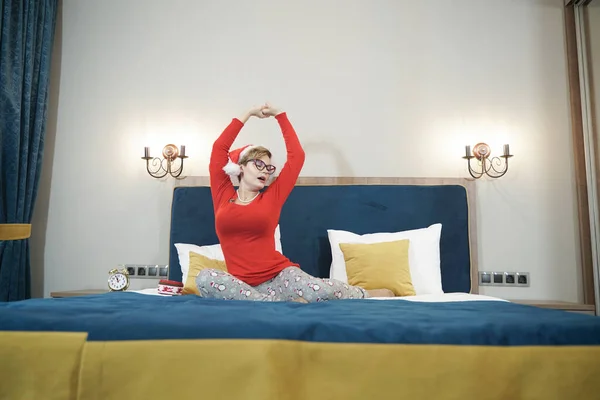 Νυσταγμένη γυναίκα με κόκκινο πουλόβερ και καπέλο santa που τεντώνεται ενώ σηκώνεται το πρωί μετά τα Χριστούγεννα στο κρεβάτι — Φωτογραφία Αρχείου