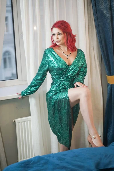 Linda mujer de pelo rojo brillante con vestido de noche de brillo de Navidad verde y de pie cerca de la ventana —  Fotos de Stock