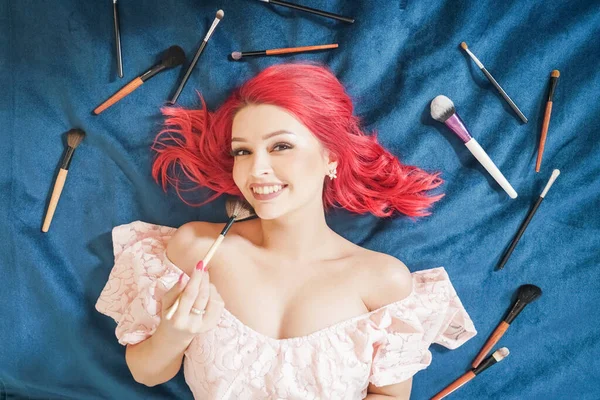 Bonita mujer con el pelo rojo acostado con maquillaje cepillos y soñando, top view girl make up master on blue background — Foto de Stock