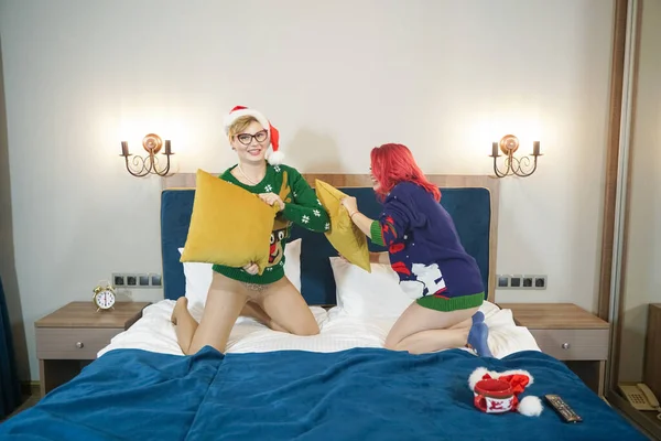 Dos lindas amigas felices tienen pelea de almohadas en suéteres de Navidad en la gran cama azul —  Fotos de Stock