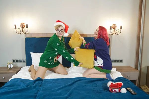 Dos lindas amigas felices tienen pelea de almohadas en suéteres de Navidad en la gran cama azul —  Fotos de Stock