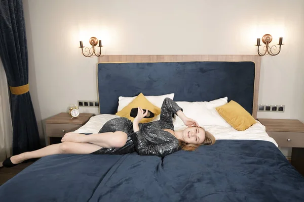Jovem mulher em elegante vestido de noite preto deitado na cama. Adulto caucasiano menina loira em roupa de lantejoulas relaxar no quarto . — Fotografia de Stock