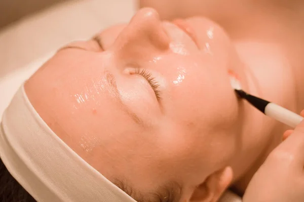 Mujer joven acostada en el salón de spa mientras hace mascarilla facial por cepillo. de cerca — Foto de Stock