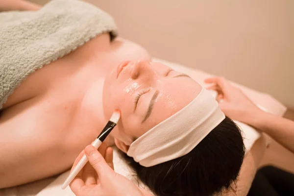 Linda mezcla de raza mujer miente y disfruta del proceso de aplicación de una máscara de cepillo en el salón de belleza en su cara —  Fotos de Stock