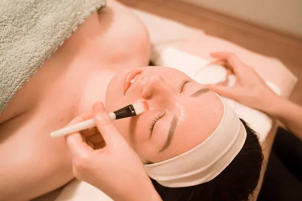 Carino mix gara donna si trova e gode il processo di applicazione di una maschera pennello nel salone di bellezza sul suo viso — Foto Stock