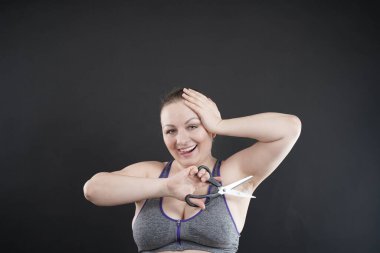 Beyaz tenli, iri yarı kadın kıllı koltuk altını gösteriyor. Kız elinde bir makas tutuyor ve neşeli, şaşkın duygular hissediyor, gülüyor. Stüdyo siyah monokrom arkaplanı