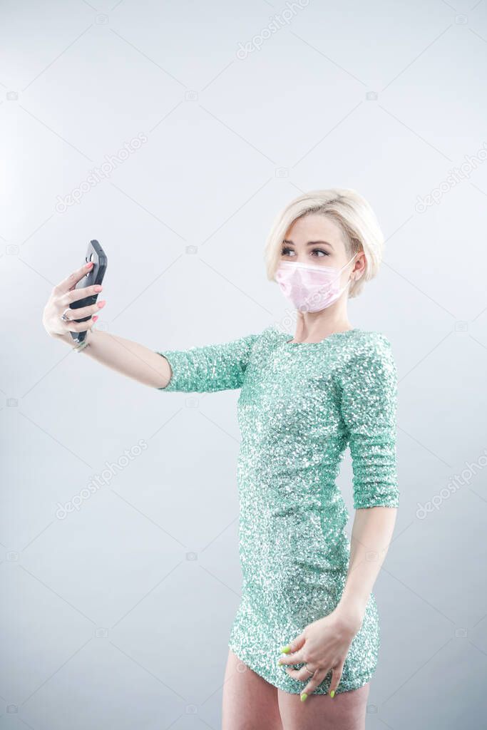 young girl in a beautiful dress and a medical mask observes the quarantine while at home alone, communicates with friends on the phone via video and is happy. woman on a white background in the Studio