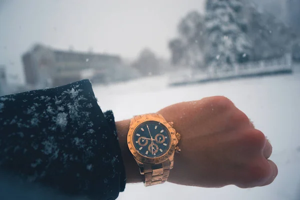 A mão de um homem com um relógio dourado contra uma queda de neve na cidade . — Fotografia de Stock