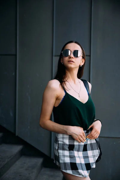 Ragazza sexy vicino a un muro scuro in una t-shirt, occhiali da sole e pantaloncini corti — Foto Stock