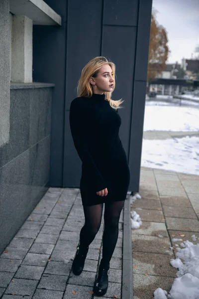 Menina em um vestido preto no inverno em um fundo de um edifício escuro — Fotografia de Stock