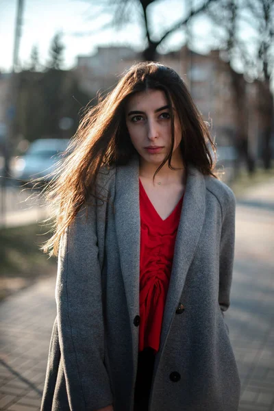 Chica en una blusa roja y un cárdigan gris sobre un fondo borroso de la ciudad a principios de primavera —  Fotos de Stock