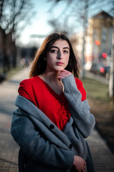 Chica en una blusa roja y un cárdigan gris sobre un fondo borroso de la ciudad a principios de primavera —  Fotos de Stock