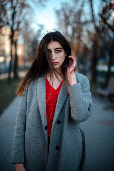 Menina em uma blusa vermelha e um casaco cinza em um fundo turvo da cidade no início da primavera — Fotografia de Stock