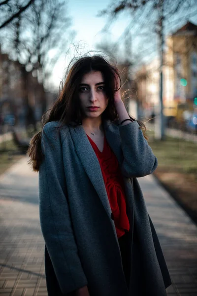 Chica en una blusa roja y un cárdigan gris sobre un fondo borroso de la ciudad a principios de primavera —  Fotos de Stock