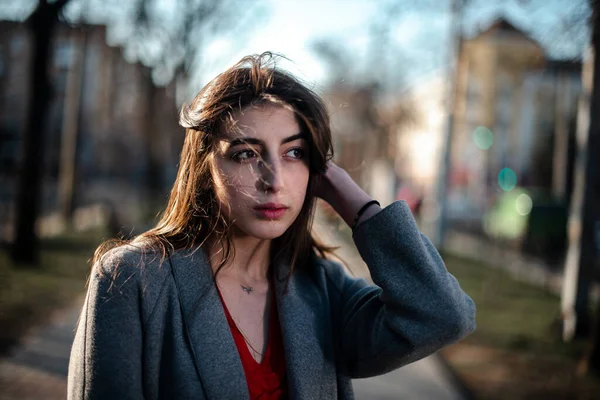 Chica en una blusa roja y un cárdigan gris sobre un fondo borroso de la ciudad a principios de primavera —  Fotos de Stock