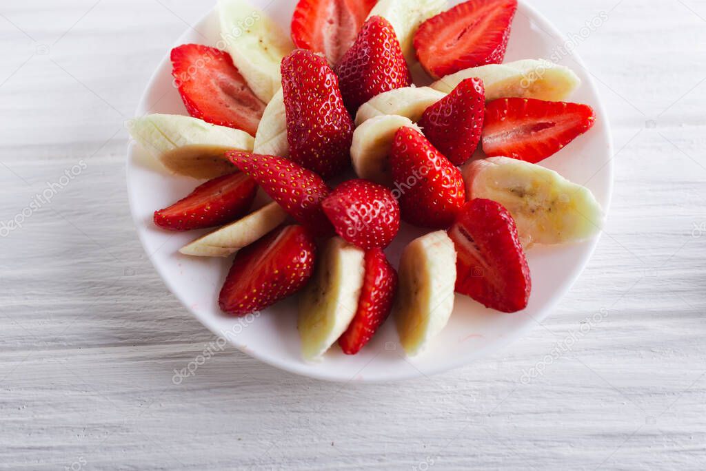 Juicy fresh sliced strawberries with banana on a white plate on a white wooden background